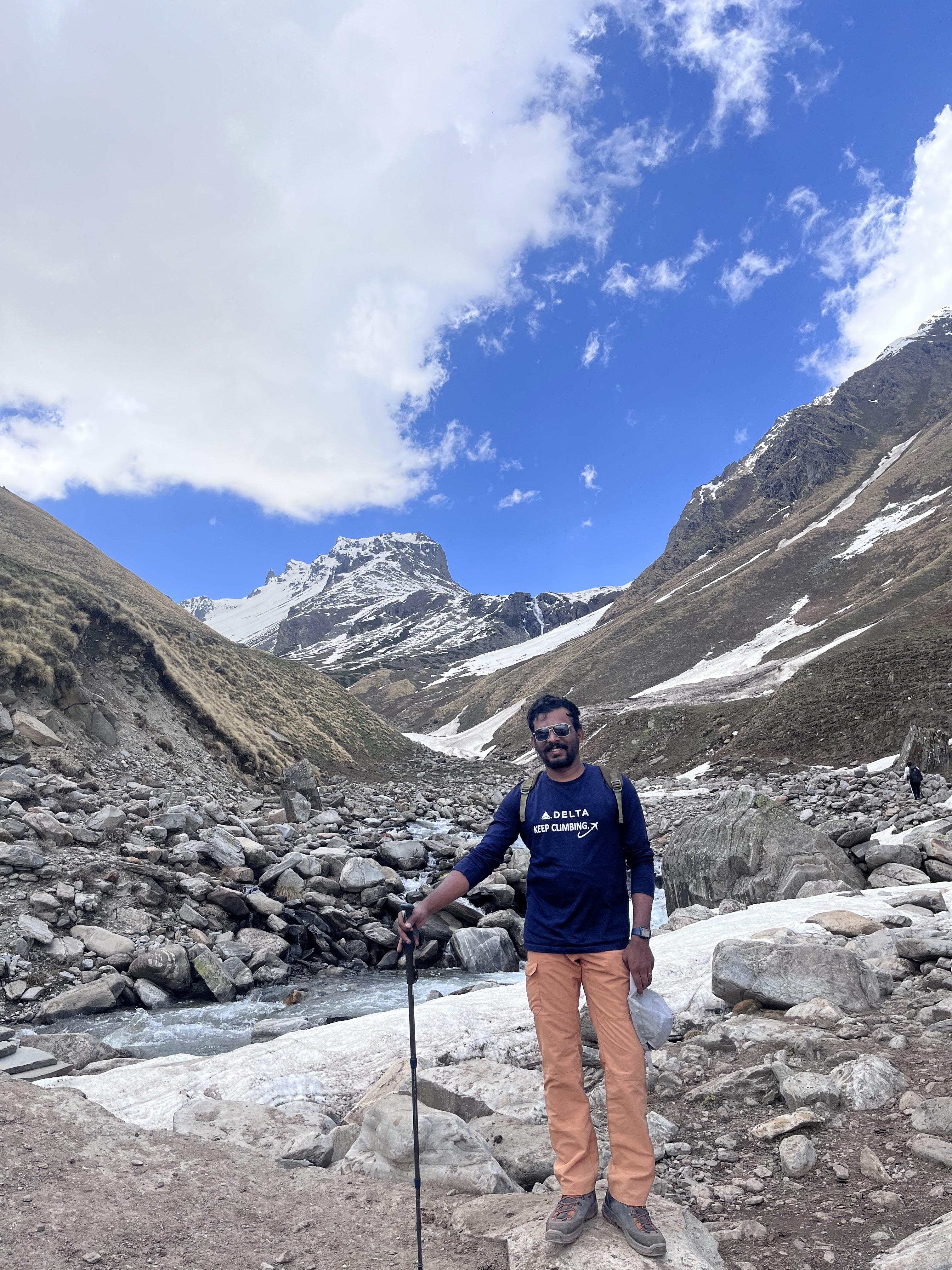 Vairav standing near a glacial stream with mountains behind him, holding a trekking pole.