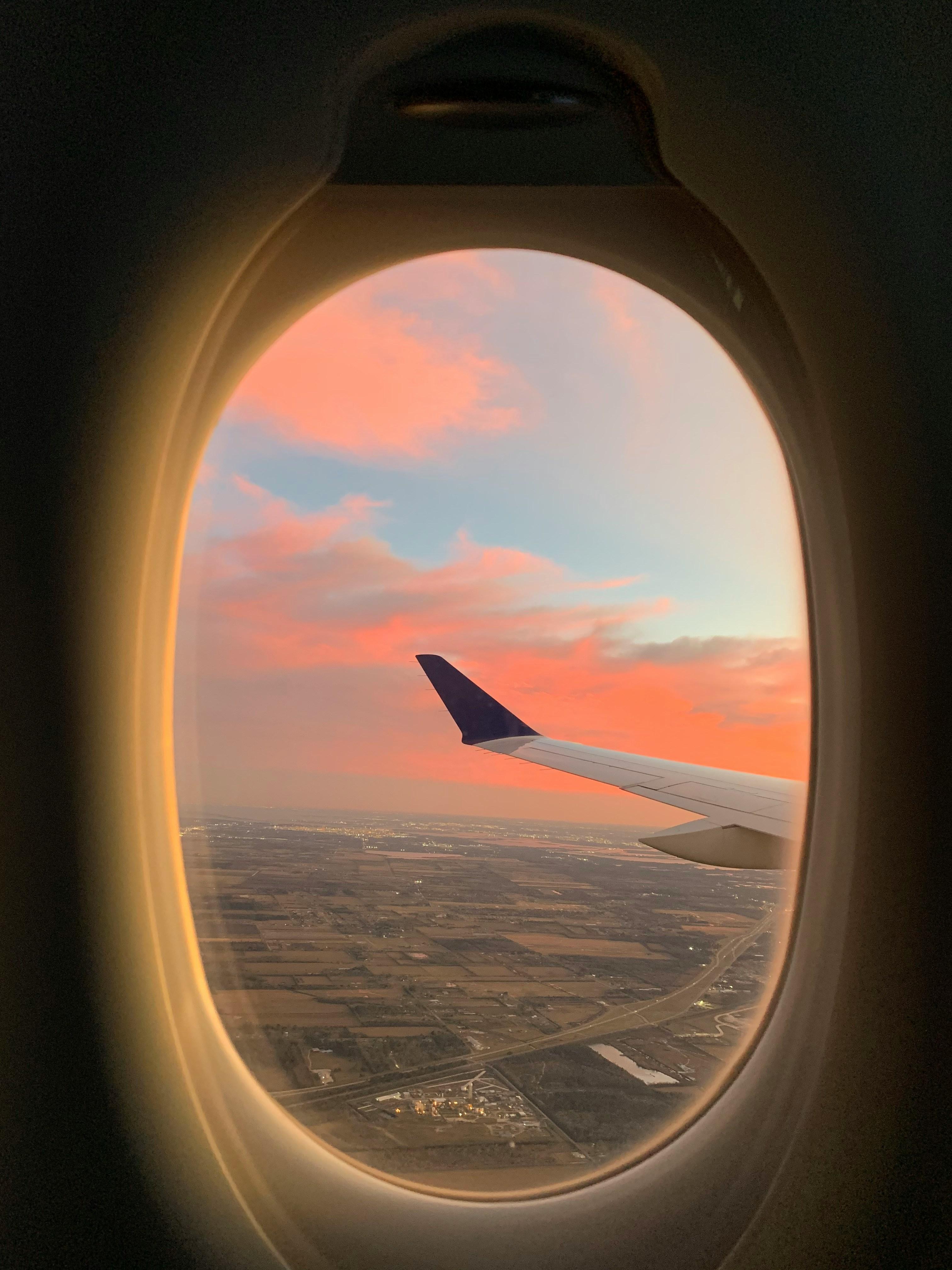 View through an airplane window at sunset, showing the wing silhouetted against an orange and pink sky above farmland.