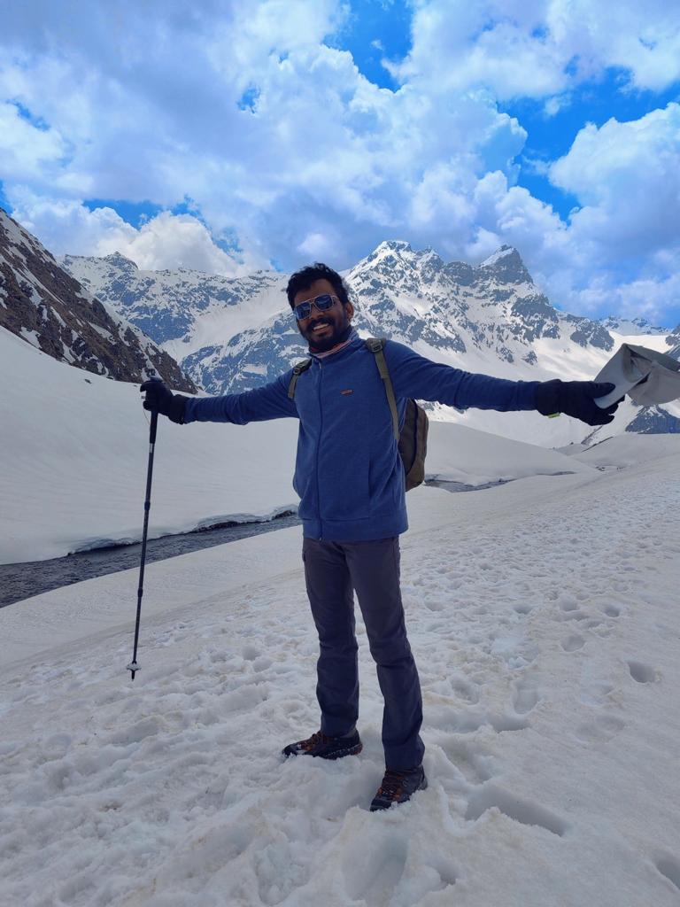 Vairav standing on a snowfield in the mountains, arms outstretched, with peaks and clouds behind him.