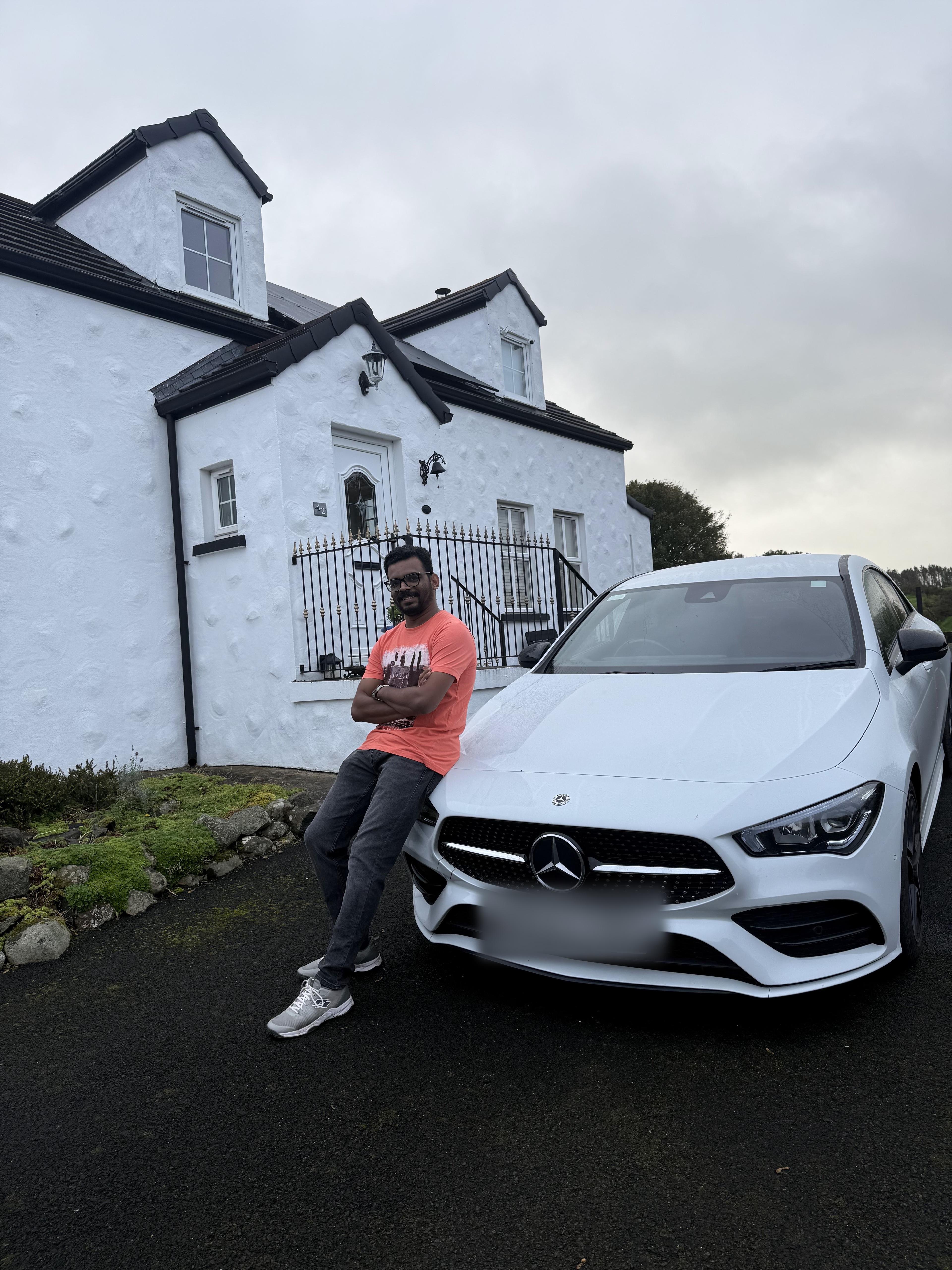 Vairav leaning against a white Mercedes parked outside a whitewashed cottage under an overcast sky.