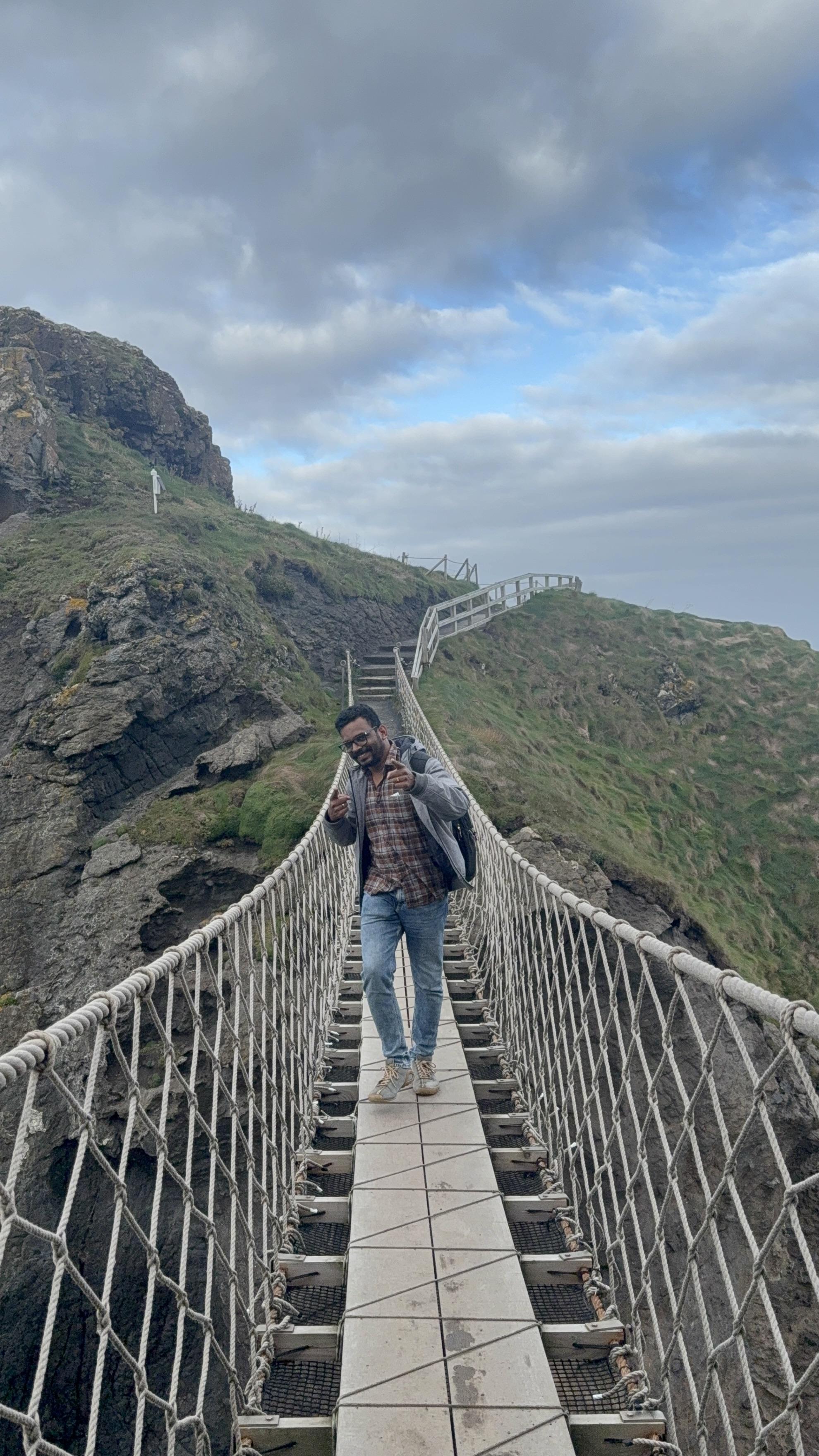 Vairav standing on a rope bridge over a cliff edge, pointing at the camera, with a grassy headland and cloudy sky behind him.