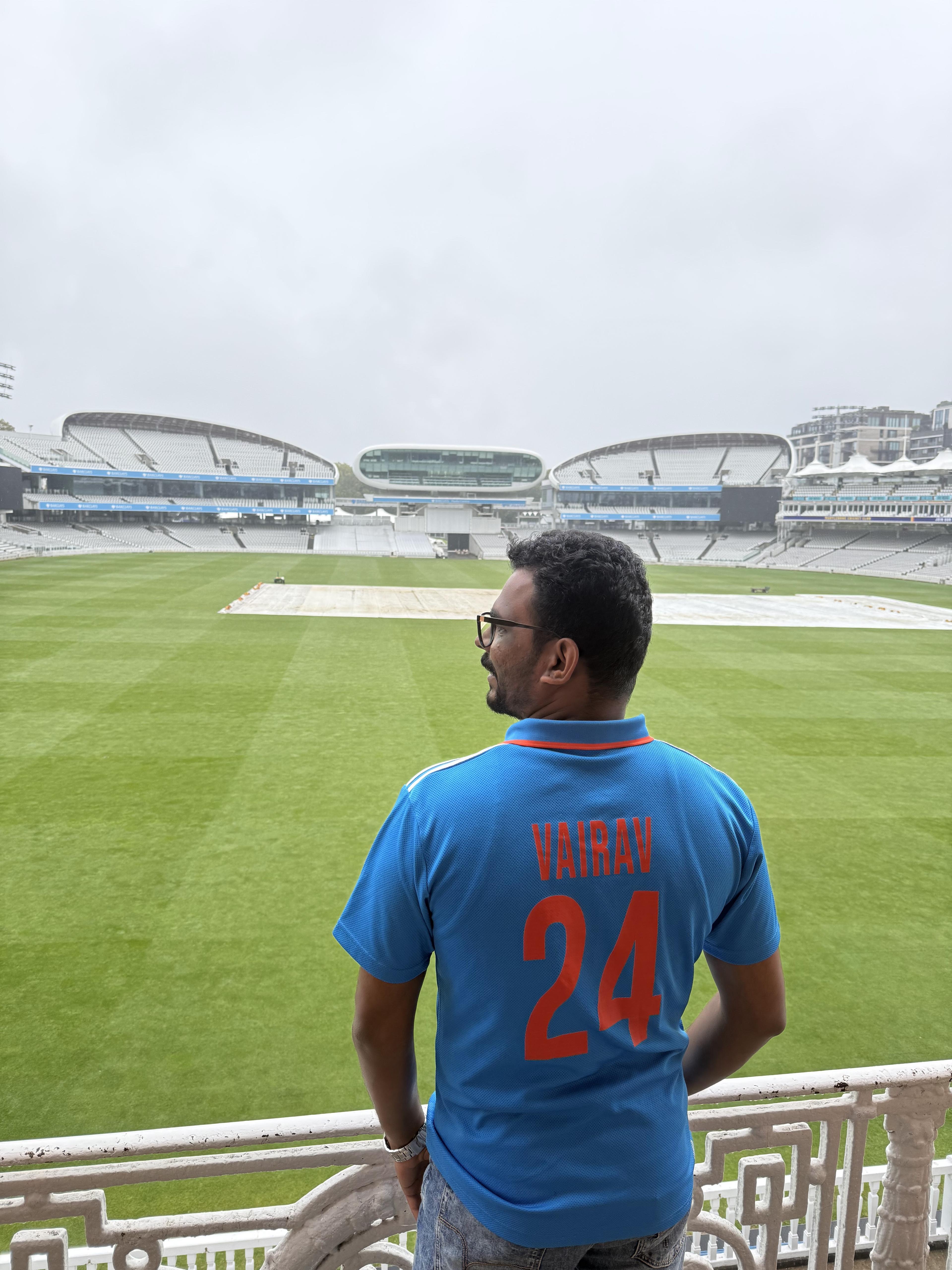 Vairav wearing an India cricket jersey printed with his name, looking out over a cricket ground from the stands.