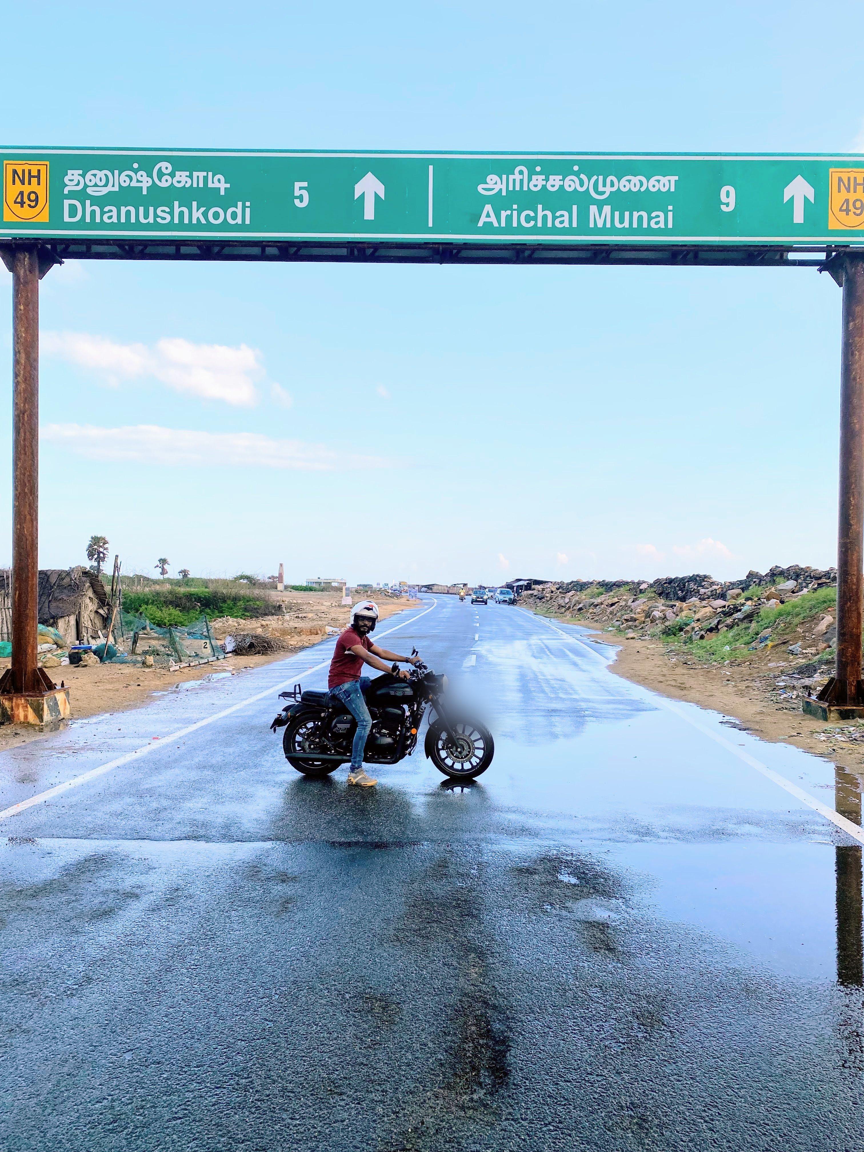 Vairav with his motorcycle on a coastal highway near Dhanushkodi, under a road sign for Dhanushkodi and Arichal Munai.
